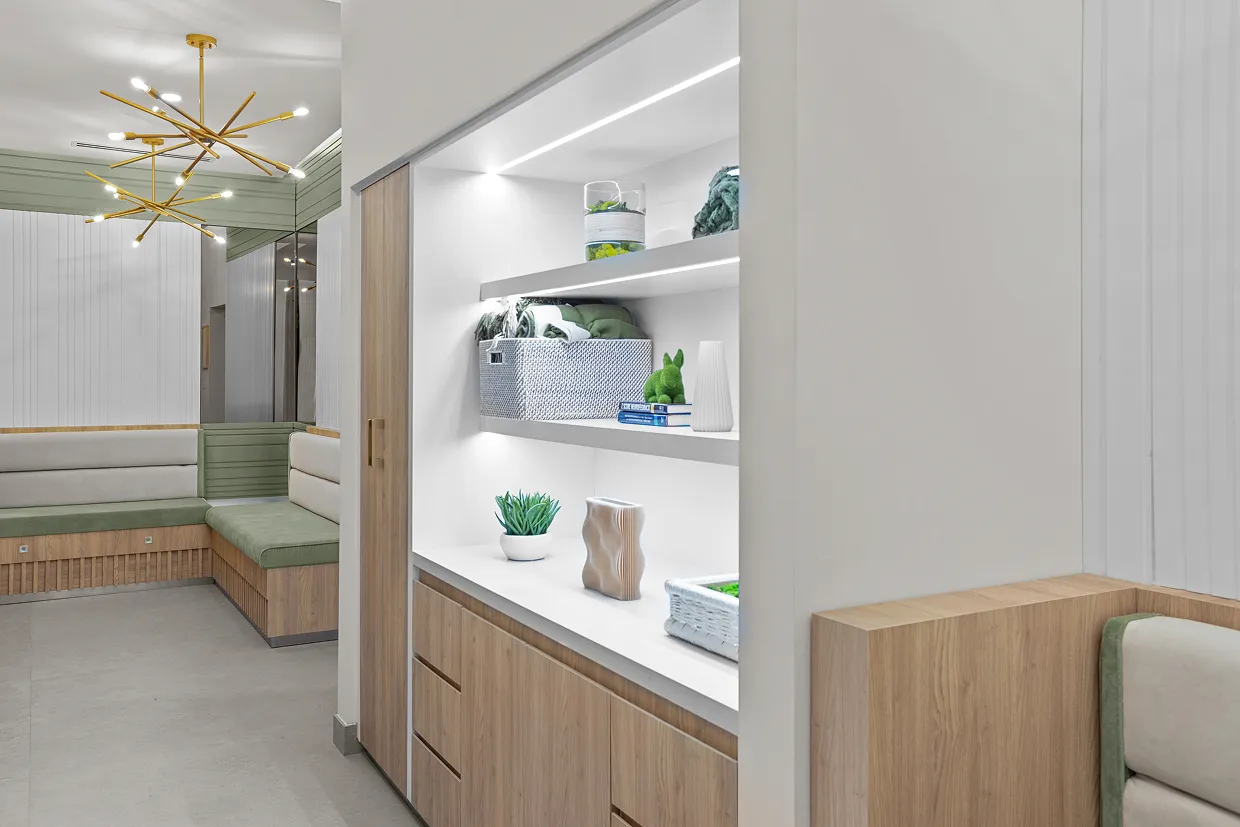 Modern waiting room with light wood and green cushioned bench seating, decorative shelves with plants and books, and a gold multi-arm chandelier.