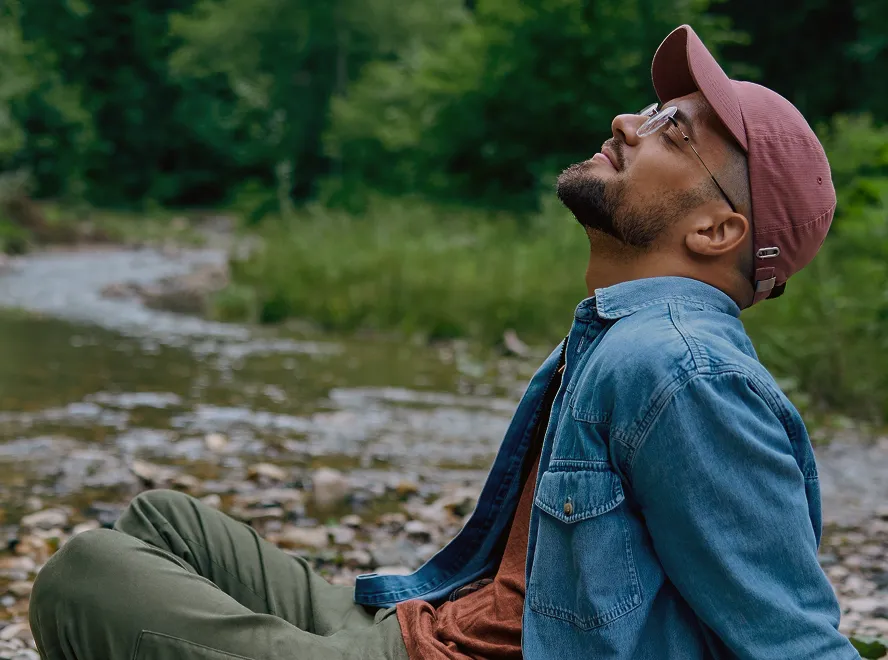 Man wearing a pink cap, glasses, denim jacket, and green pants sitting by a riverbank with greenery in the background.