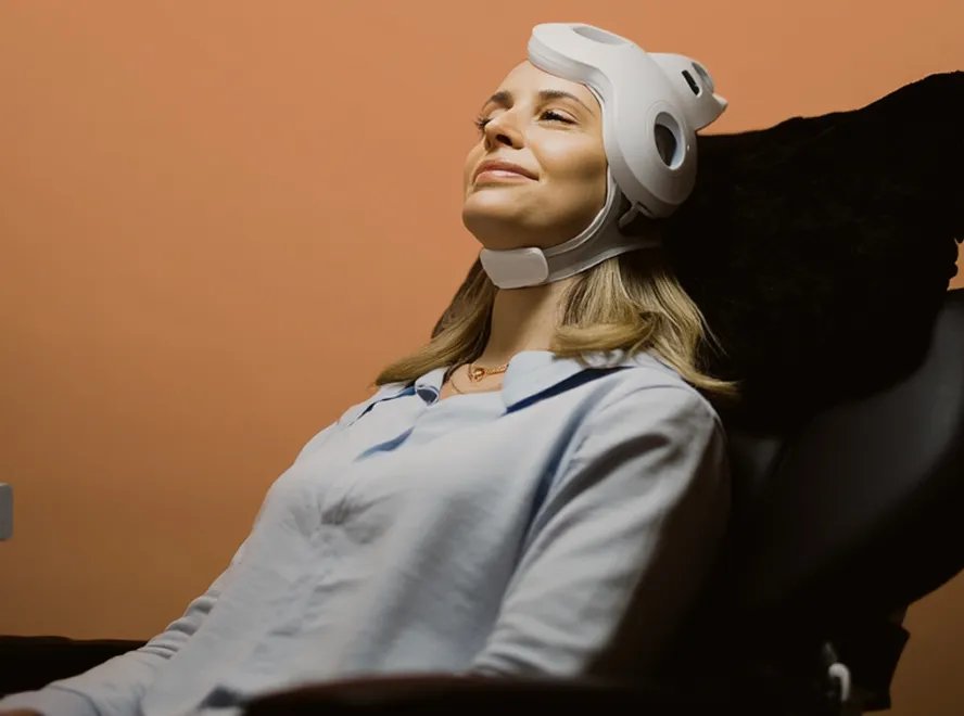 A woman wearing a white medical headgear reclines on a black chair against a brown background, appearing calm and content.