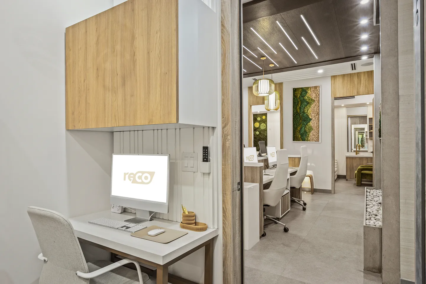 Modern office workspace with a white desk, computer, and ergonomic chair next to a hallway with white chairs, wooden cabinets, vertical garden art, and ceiling lights.