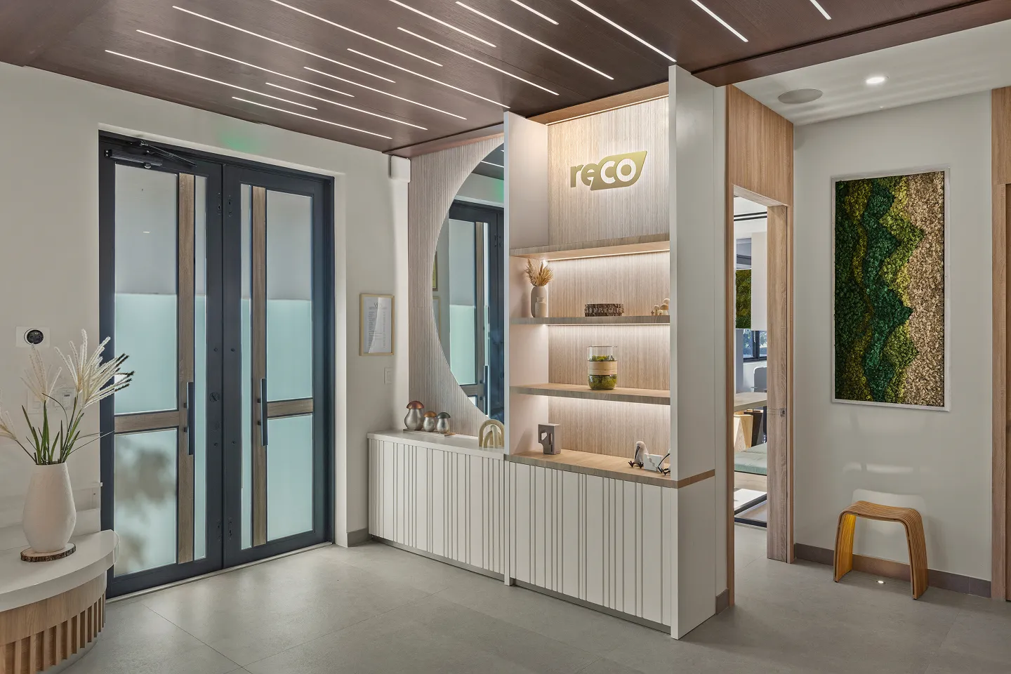 Modern reception area with frosted glass double doors, wooden paneled ceiling with linear lights, illuminated shelves displaying decor, and a moss wall art beside a wooden stool.