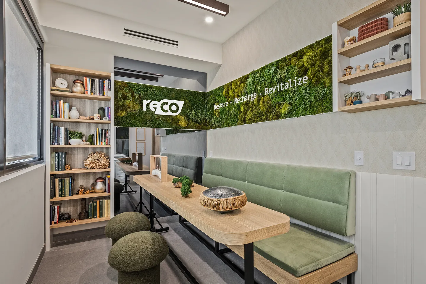 Modern meeting room with green cushioned bench, wooden table, mushroom-shaped stools, and a moss wall display with the words 'reco Restore • Recharge • Revitalize'.