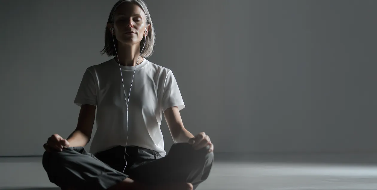 Woman sitting cross-legged in a dimly lit room meditating with eyes closed and wearing earphones.