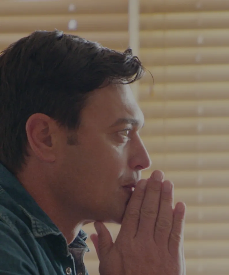 Side profile of a man with dark hair looking thoughtful with his hands pressed together near his mouth.