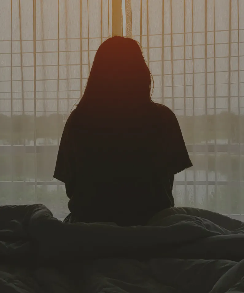 Silhouette of a person sitting on a bed facing a window with sheer curtains during daytime.