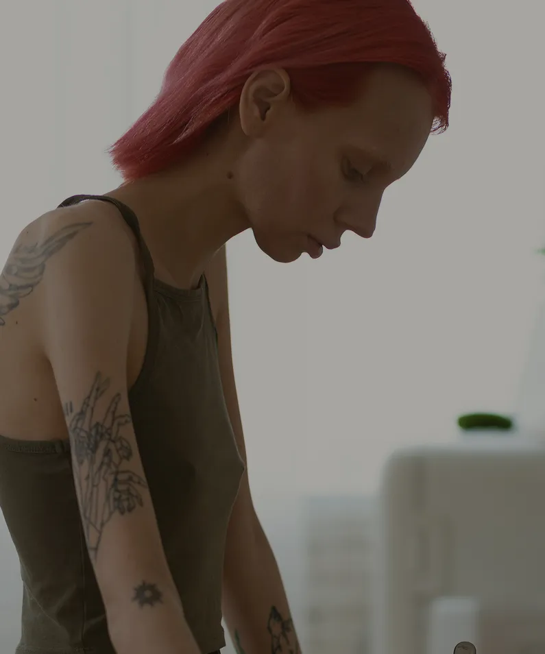 Person with red hair and tattoos on their arms wearing a sleeveless dark top, leaning forward.